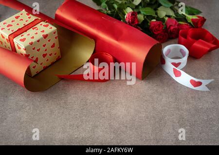 Boîte-cadeau enveloppé dans du papier rouge, roses et des matériaux d'emballage sur fond brun. Copy space Banque D'Images