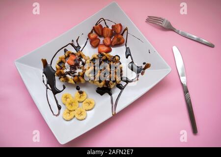 Gaufres avec du sirop de chocolat, fraise et banane sur une plaque sur un fond rose Banque D'Images