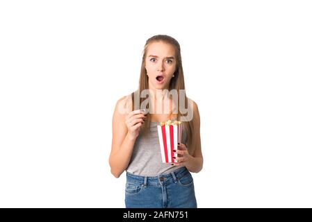 Fille mange du pop-corn et s'étonne à l'appareil photo. Une belle blonde montre l'émotion de surprise tout en regardant un film. Fille isolée. Banque D'Images