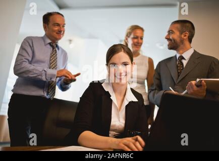 Portrait of businesswoman sitting at desk with laptop, en tant que co-travailleurs se tenir derrière elle. Banque D'Images