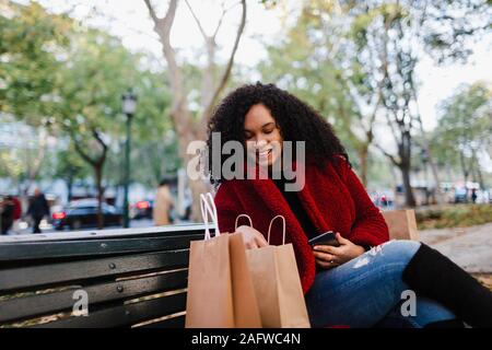 Smiling young woman looking in shopping bags on park bench Banque D'Images
