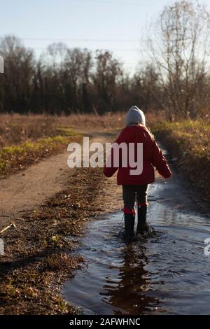 3 ans enfant s'éclabousser dans une flaque avec Red Jacket et wellies sur une journée d'hiver ensoleillée. Vue arrière, Vertical shot. Banque D'Images