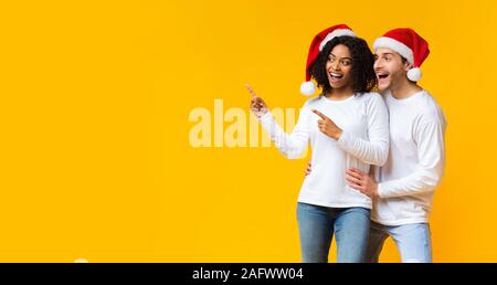 Cheerful couple interracial dans santa hats pointant de côté à copy space Banque D'Images