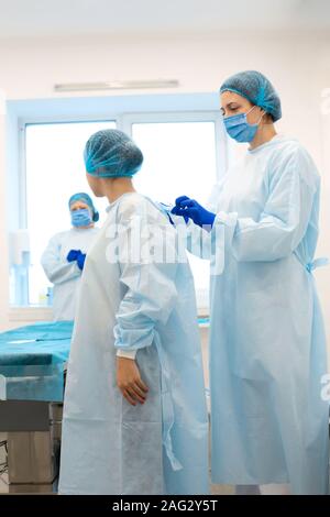 Une infirmière robes une chirurgien dans un costume stérile avant la chirurgie. Banque D'Images