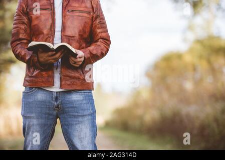 Homme portant une veste en cuir debout sur une route vide et la lecture de la bible avec un arrière-plan flou Banque D'Images