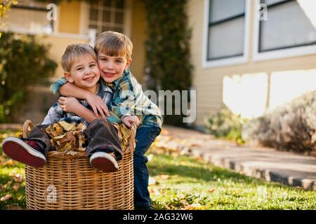 Boy hugging son petit frère assis dans le panier plein de feuilles en automne. Banque D'Images