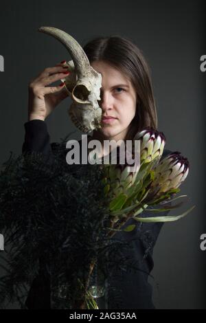 Portrait d'une jeune fille brune se tenant la moitié d'un crâne de chèvre en face de son visage et un bouquet de fleurs noires et les asperges, duplicité concept Banque D'Images