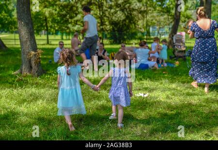 Deux filles d'âge préscolaire dans des robes turquoise et violette marchant et tenant les mains dans le parc tout en approchant un groupe de personnes ayant pique-nique dans la nature Banque D'Images