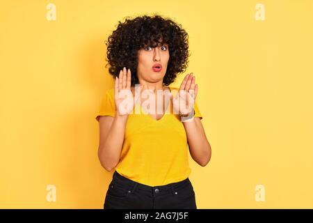 Jeune femme arabe avec des cheveux bouclés vêtus de t-shirt debout sur fond jaune isolé éloigne les mains paumes montrant refus et déni avec afra Banque D'Images
