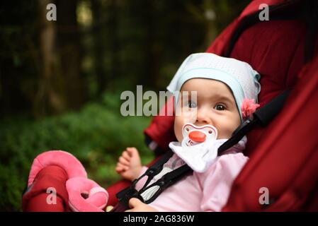 Adorable baby girl à l'extérieur en rouge en poussette forêt. Sucette nouveau-né avec fond vert Banque D'Images