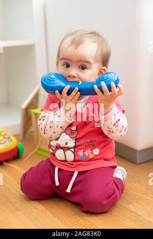 Caucasien mignon bébé de 1 an fille mord téléphone bleu jouet, concept de la dentition, les morsures de l'enfant des jouets. Fille en chemise rouge hello kitty Banque D'Images