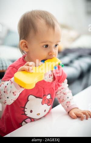 Caucasien mignon bébé de 1 an fille jouet guitare jaune morsures, concept de jouets de dentition, les morsures de l'enfant. Fille en chemise rouge hello kitty Banque D'Images