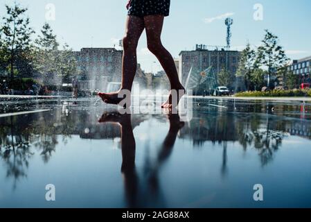 Reflet d'une personne pieds nus sur le trottoir humide avec fontaines d'eau en arrière-plan Banque D'Images
