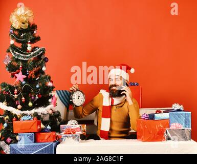 Santa est titulaire cellulaire et un réveil. Célébration des gadgets et concept. Homme avec barbe et heureux face envoie des vœux pour le Nouvel An. Le Père Noël parle sur téléphone mobile près de sapin sur fond rouge. Banque D'Images