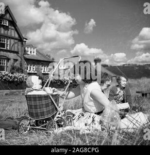 Années 1960, historique, une mère et un enfant en bas âge assis dans un petit buggy ou poussette avec un siège tartan, assis à l'extérieur sur une zone herbeuse quelques maisons, Angleterre, Royaume-Uni. Banque D'Images