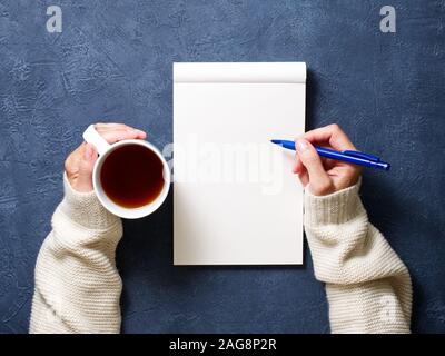 Femme écrit dans l'ordinateur portable sur une table, bleu foncé dans la main tenant une chemise crayon, tasse de thé, sketchbook dessin, vue de dessus Banque D'Images