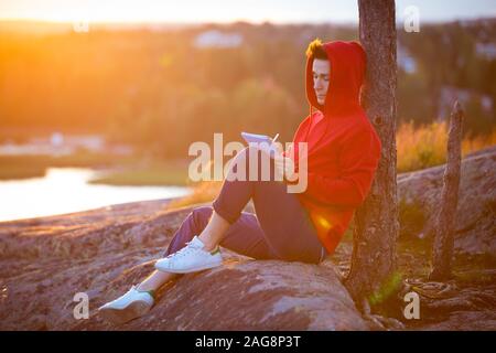 Jeune homme à hoodie assis sur le dessus de la roche, écrivant dans l'ordinateur portable. Beau paysage de coucher de soleil. L'homme s'inspire de la nature. Finlande, Espoo Banque D'Images