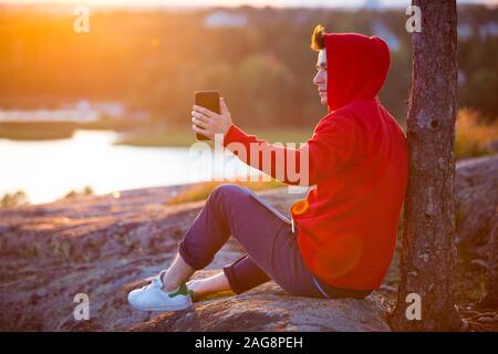 Jeune homme à hoodie assis sur le dessus de la roche, prendre une photo au téléphone. Beau paysage de coucher de soleil. L'homme s'inspire de la nature. Finlande, Espoo Banque D'Images