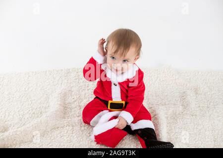 Stock studio photo avec fond blanc d'un bébé déguisé en père Noël avec la main sur le visage entouré par des jouets Banque D'Images