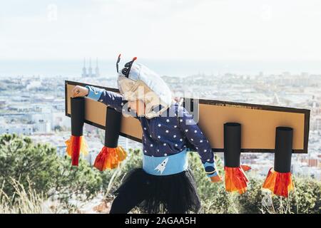 Drôle de petit enfant fille joue avec des super-héros, de costumes ailes avion en carton et casque d'astronaute comme voler en face d'un paysage urbain, imagina Banque D'Images