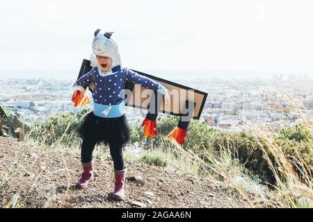 Drôle de petit enfant fille voler comme un super héros avec des ailes d'avion en carton, costume et casque d'astronaute en face d'un paysage urbain, de l'imagination Banque D'Images