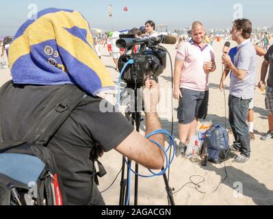 Interview à la télévision, Rio, au Brésil. Un opérateur de l'appareil photo lui-même les protections contre le soleil comme il filme un emplacement plat interview sur une plage de Rio. Banque D'Images