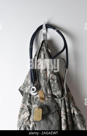 Concept de santé militaire : un ensemble de dog tags, stéthoscope et camouflage veste suspendue à un crochet sur un mur blanc. Banque D'Images