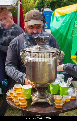 Adygea, Russie - le 21 septembre 2019 : un homme barbu se trouve à la table avec un samovar et pots de miel et prépare le thé pour les touristes au festival de Banque D'Images