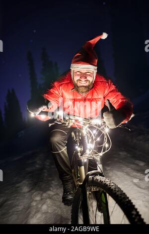 Portrait d'homme barbu en veste rouge et Noël hat le vélo à l'hiver la forêt enneigée sous ciel de nuit avec des étoiles Banque D'Images