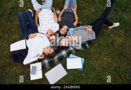 En tenant à l'aide de selfies smartphone. Groupe de jeunes étudiants dans les tenues sur l'herbe verte dans la journée Banque D'Images