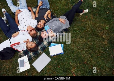 En tenant à l'aide de selfies smartphone. Groupe de jeunes étudiants dans les tenues sur l'herbe verte dans la journée Banque D'Images