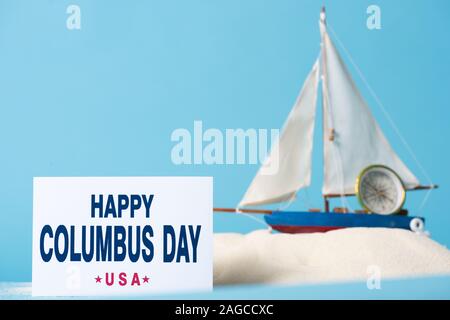 Carte avec happy Columbus Day inscription près de bateau miniature dans le sable blanc sur bleu isolé Banque D'Images