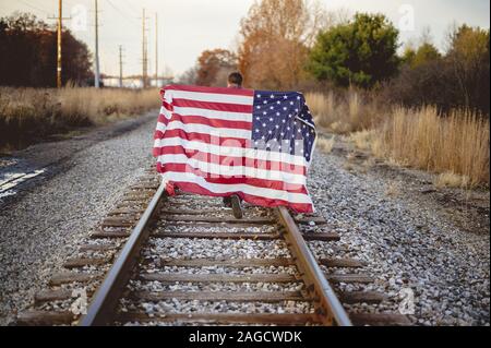 Homme tenant le drapeau des États-unis derrière lui et marchant sur les voies ferrées Banque D'Images