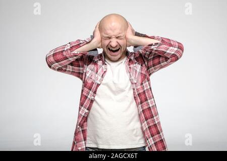 L'homme non rasé chauve, dans le stress, il a couvert ses oreilles avec les mains afin de ne pas entendre des sons ennuyeux lui et crie fort. Banque D'Images