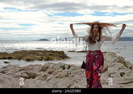 Jeune femme à la mode hippie avec de longs cheveux bruns et un pantalon évasé floral pose par la mer sur les rochers en dessous d'une moue, Cloudscape et jouer wit Banque D'Images