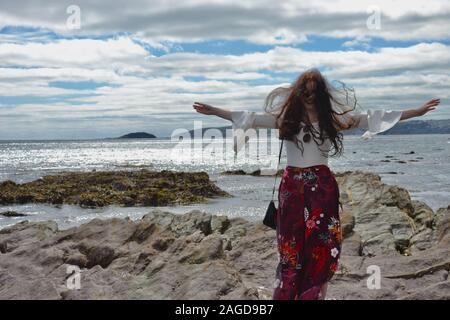 Jeune femme à la mode hippie avec de longs cheveux bruns et un pantalon évasé floral pose par la mer sur les rochers en dessous d'une propagation d'armes, Cloudscape et malpropre h Banque D'Images