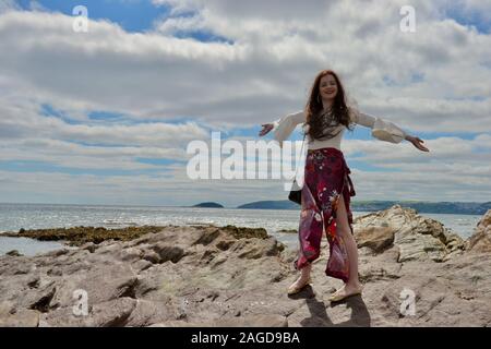 Jeune femme à la mode hippie avec de longs cheveux bruns et un pantalon évasé floral pose par la mer sur des rochers sous un bras et cloudscape, smiling Banque D'Images