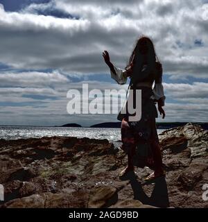 Jeune femme à la mode hippie avec de longs cheveux bruns et un pantalon évasé floral pose par la mer sur des rochers sous un cloudscape ; et rétroéclairé avec son ha Banque D'Images