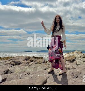 Jeune femme à la mode hippie avec de longs cheveux bruns et un pantalon évasé floral pose par la mer sur des rochers sous un cloudscape, rire avec sa main Banque D'Images
