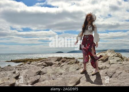 Jeune femme à la mode hippie avec de longs cheveux bruns et un pantalon évasé floral pose par la mer sur des rochers sous un cloudscape, souriant et avec windsw Banque D'Images