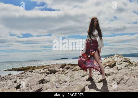 Jeune femme à la mode hippie avec de longs cheveux bruns et un pantalon évasé floral pose par la mer sur des rochers sous un cloudscape Banque D'Images