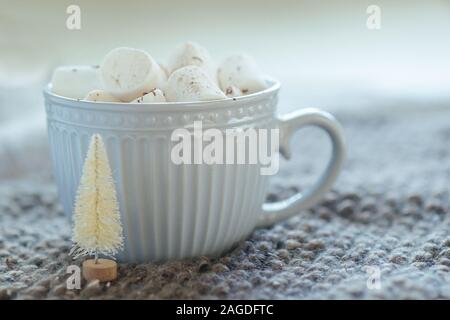 Petit arbre de Noël blanc artificiel à côté de grande tasse de café ou de chocolat chaud avec des guimauves sur table avec des nappes de gros tricot. Banque D'Images