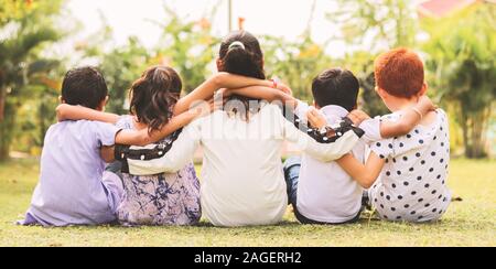 Vue arrière, Groupe d'amis les enfants de la maternelle ethnique multi bras autour de assis ensemble à l'extérieur du parc - Concept montrant de l'amitié de la petite enfance Banque D'Images