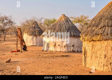 Trois africains traditionnels, des maisons aux toits de chaume dans un village tribal Himba en Namibie rurale, tandis que les vêtements pendent des vieux arbres. Banque D'Images