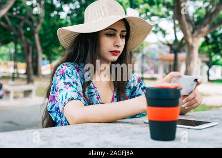 Tasse réutilisable en premier plan. Arrière-plan flou Indian female en extérieur parc holding mobile phone. Elle ne pouvait être vu travailler hors du bureau, vidéo Banque D'Images