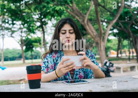 Une femme indienne moderne en extérieur parc holding mobile phone. Sur la table est une tasse réutilisable respectueux de l'environnement, et d'un écran tactile Tablet dev Banque D'Images