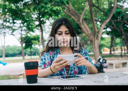 Une femme indienne moderne en extérieur parc holding mobile phone. Sur la table est une tasse réutilisable respectueux de l'environnement, et d'un écran tactile Tablet dev Banque D'Images