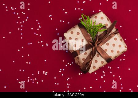 Bricolage cadeau dans du papier craft et avec un brun satiné ruban sur fond rouge et argent confettis. Le concept d'offrir des cadeaux, des surprises, de Noël et de N Banque D'Images