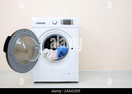Machine à laver moderne avec des tas de linge dans le tambour et ouvrez la porte se dresse sur plancher en bois près de mur beige Banque D'Images