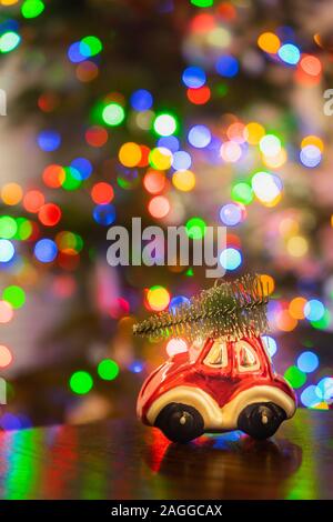 Voiture miniature ornement rouge transportant arbre de Noël sur table en bois avec des lumières de Noël colorées en arrière-plan flou Banque D'Images